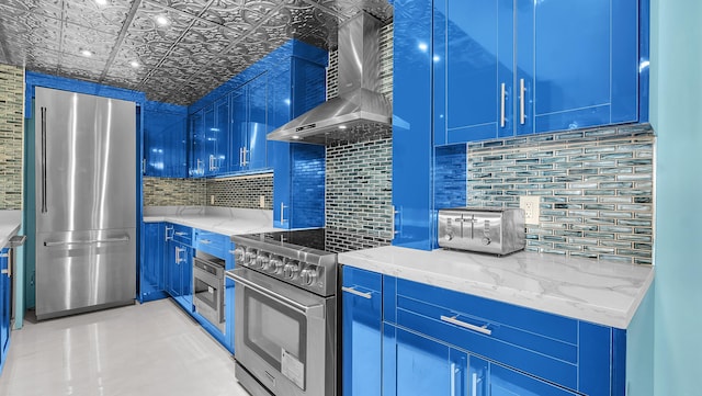 kitchen featuring tasteful backsplash, blue cabinets, stainless steel appliances, light stone countertops, and wall chimney range hood