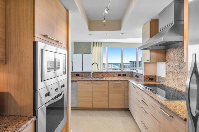 kitchen featuring wall chimney exhaust hood, appliances with stainless steel finishes, light stone counters, light brown cabinets, and a sink