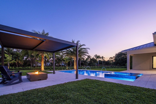 pool at dusk with a lawn, a patio, an outdoor fire pit, pool water feature, and an in ground hot tub