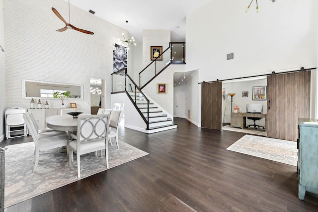 dining space with dark wood-style floors, stairs, a towering ceiling, a barn door, and ceiling fan with notable chandelier