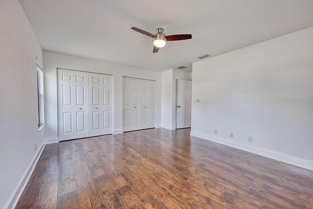 unfurnished bedroom with baseboards, visible vents, dark wood finished floors, ceiling fan, and two closets