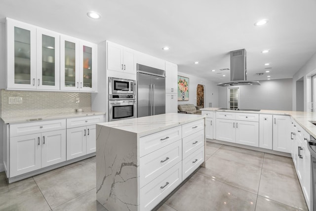 kitchen featuring island exhaust hood, light tile patterned floors, backsplash, glass insert cabinets, and built in appliances