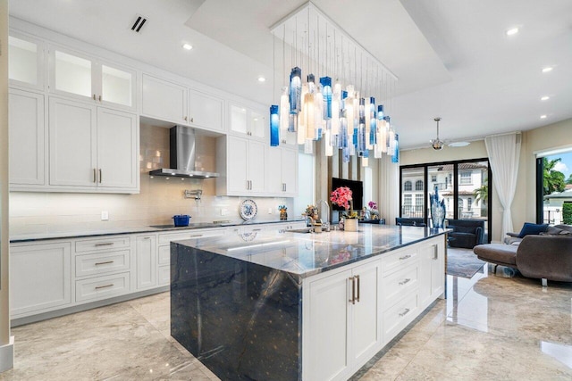 kitchen featuring white cabinets, glass insert cabinets, a large island with sink, wall chimney range hood, and pendant lighting