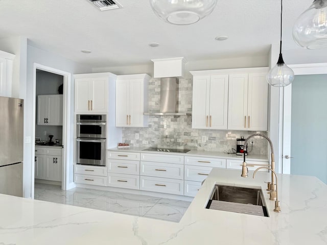 kitchen featuring white cabinets, wall chimney exhaust hood, appliances with stainless steel finishes, hanging light fixtures, and a sink