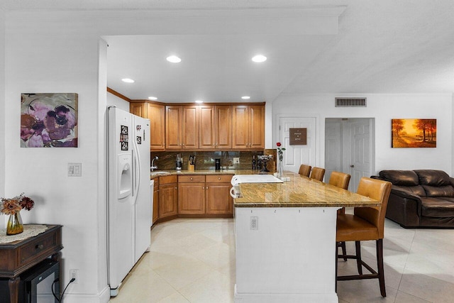 kitchen featuring visible vents, a kitchen bar, brown cabinets, white refrigerator with ice dispenser, and decorative backsplash