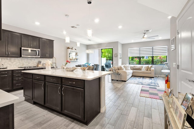 kitchen with a center island with sink, light hardwood / wood-style flooring, hanging light fixtures, dark brown cabinets, and decorative backsplash