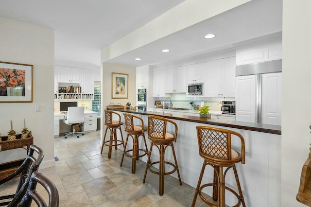 kitchen featuring kitchen peninsula, built in desk, built in appliances, white cabinets, and a kitchen bar