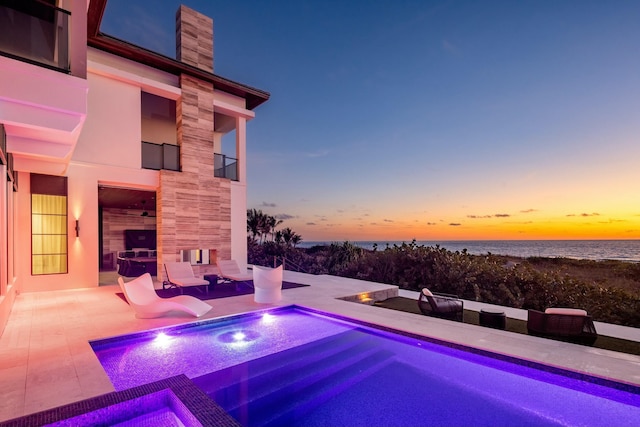 pool at dusk with a water view, an in ground hot tub, a tiled fireplace, and a patio area