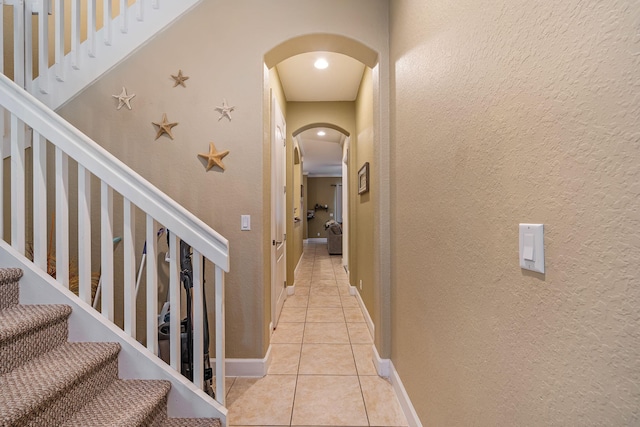 hallway featuring arched walkways, a textured wall, stairway, light tile patterned flooring, and baseboards