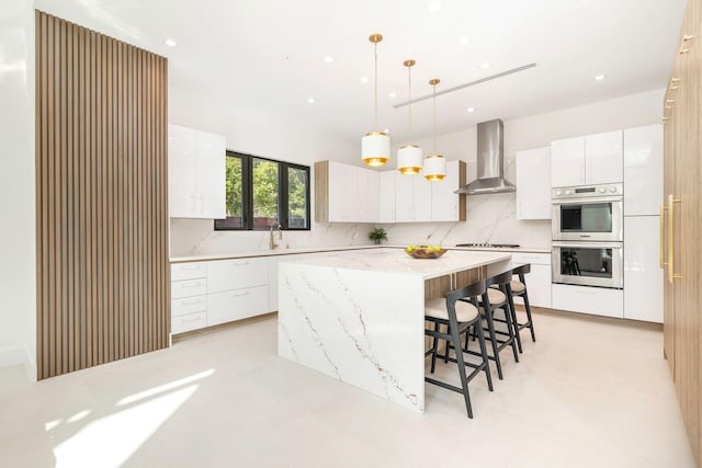 kitchen featuring a kitchen island, decorative light fixtures, white cabinets, stainless steel double oven, and wall chimney exhaust hood