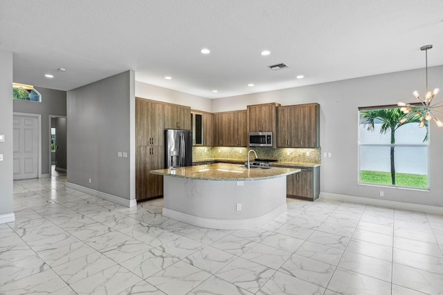 kitchen with hanging light fixtures, appliances with stainless steel finishes, an island with sink, light stone countertops, and backsplash