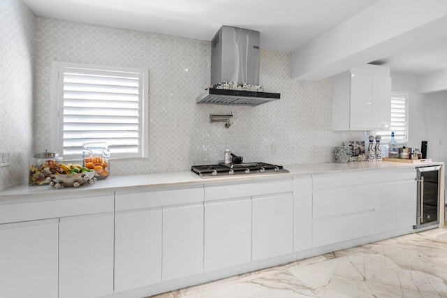 kitchen featuring a healthy amount of sunlight, wine cooler, stainless steel gas stovetop, white cabinetry, and wall chimney range hood