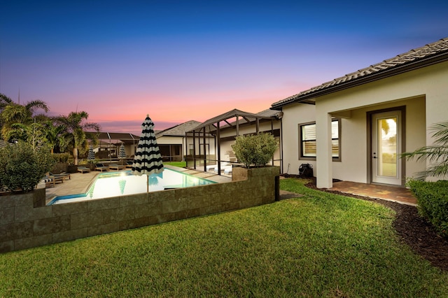 pool at dusk with an outdoor pool, glass enclosure, a lawn, and a patio