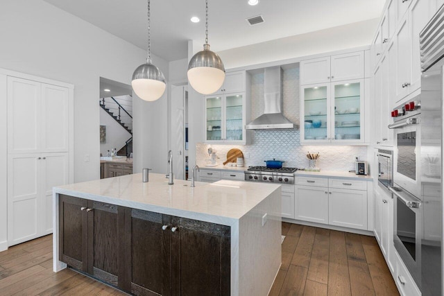 kitchen featuring stainless steel gas stovetop, an island with sink, white cabinets, light stone counters, and wall chimney range hood