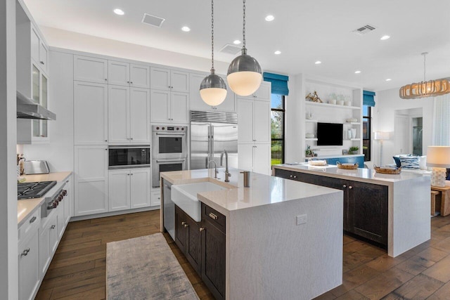 kitchen with a kitchen island with sink, built in appliances, sink, and hanging light fixtures