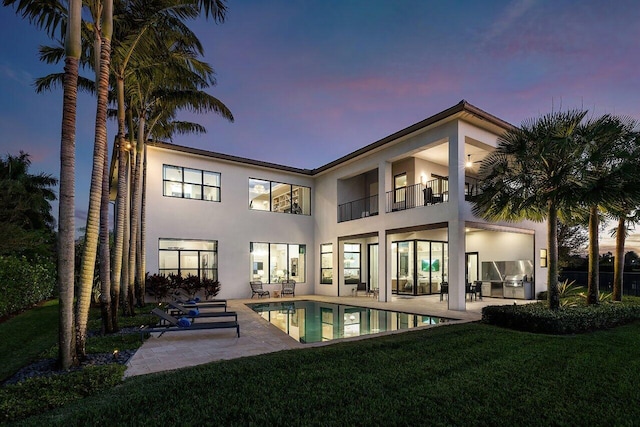 back house at dusk with a patio, a balcony, and a lawn