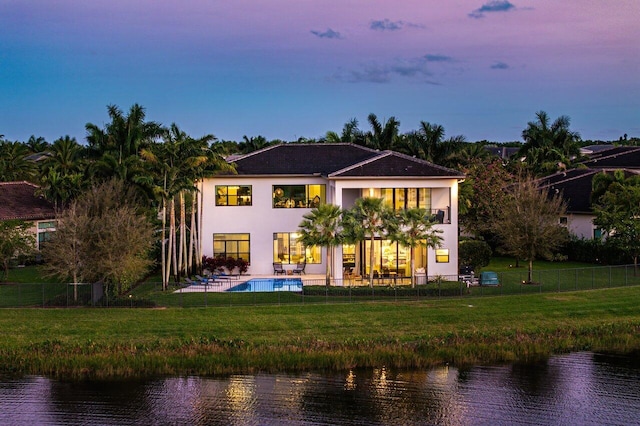 back house at dusk with a water view, a balcony, a lawn, a fenced in pool, and a patio