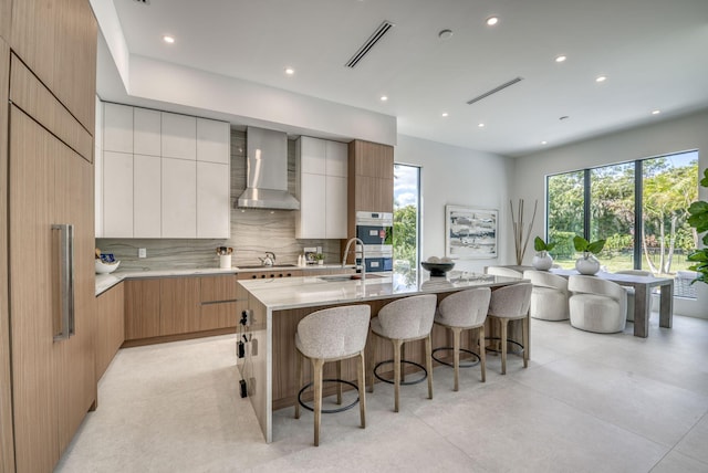 kitchen with wall chimney range hood, sink, white cabinetry, backsplash, and a large island with sink