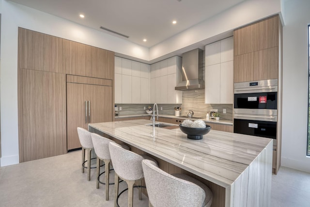 kitchen with a large island, wall chimney range hood, sink, a breakfast bar area, and paneled built in refrigerator