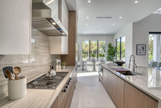 kitchen featuring light tile patterned flooring, wall chimney exhaust hood, sink, light stone countertops, and backsplash