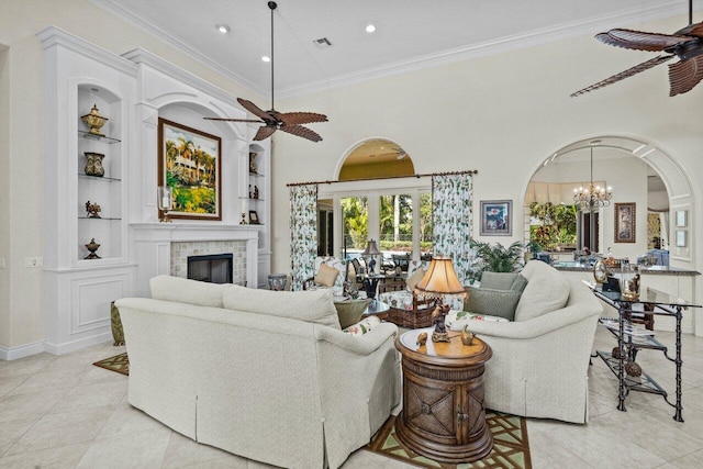 tiled living room with crown molding, a tile fireplace, a towering ceiling, built in shelves, and ceiling fan with notable chandelier