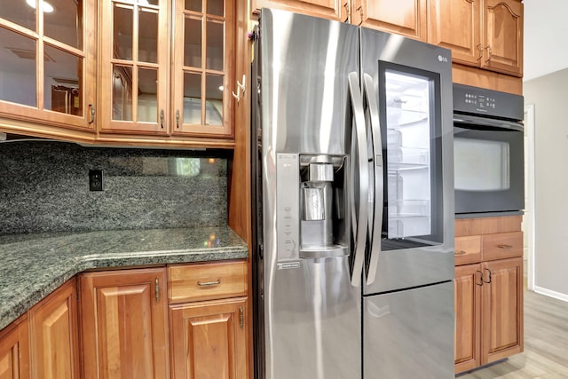 kitchen featuring stainless steel fridge with ice dispenser, light hardwood / wood-style flooring, dark stone countertops, oven, and backsplash