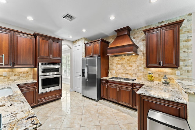 kitchen featuring light tile patterned floors, appliances with stainless steel finishes, light stone counters, tasteful backsplash, and custom range hood