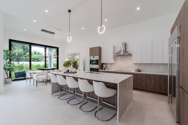 kitchen with pendant lighting, white cabinets, a kitchen island with sink, stainless steel appliances, and wall chimney range hood