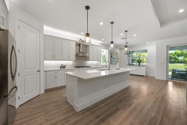 kitchen with stainless steel refrigerator, an island with sink, white cabinets, decorative light fixtures, and wall chimney exhaust hood