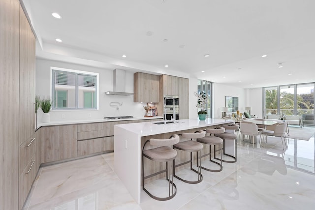 kitchen featuring an island with sink, sink, a kitchen bar, stainless steel gas cooktop, and wall chimney range hood