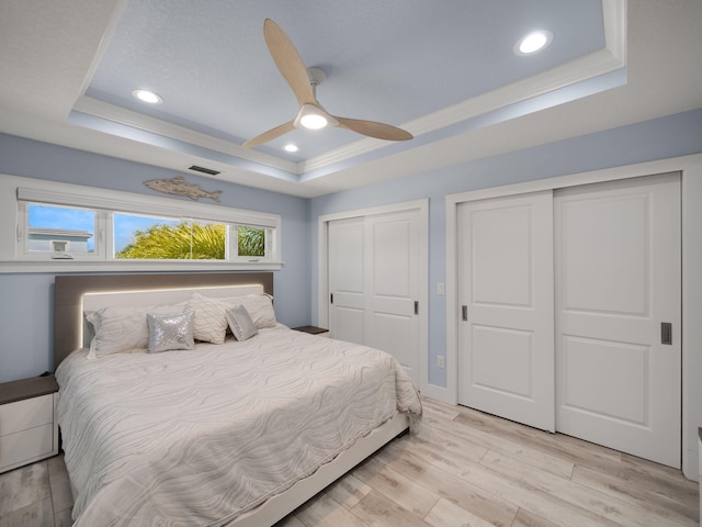 bedroom featuring multiple closets, ceiling fan, a raised ceiling, crown molding, and light wood-type flooring