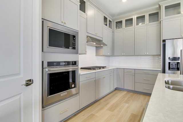 kitchen featuring light wood finished floors, stainless steel appliances, light countertops, glass insert cabinets, and under cabinet range hood