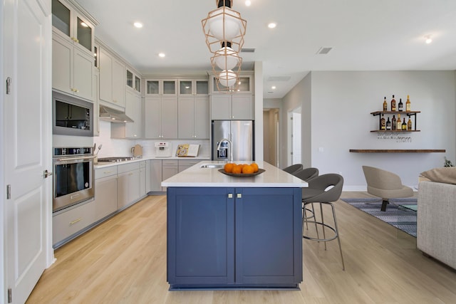 kitchen featuring pendant lighting, stainless steel appliances, light countertops, glass insert cabinets, and a kitchen island with sink