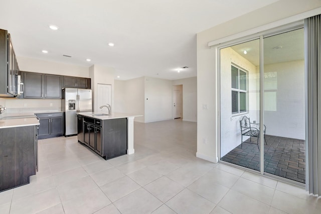 kitchen with light tile patterned floors, stainless steel fridge, a center island with sink, light countertops, and a sink