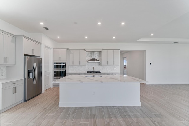 kitchen with decorative backsplash, wall chimney range hood, light stone counters, stainless steel appliances, and a center island with sink