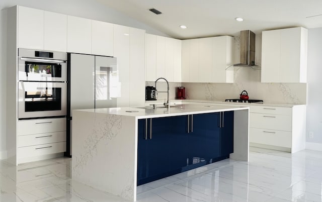 kitchen featuring double oven, white cabinetry, an island with sink, sink, and wall chimney range hood