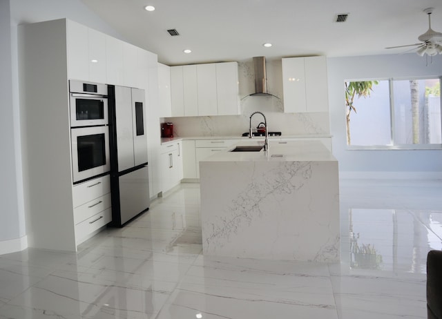 kitchen featuring wall chimney range hood, stainless steel refrigerator, double oven, tasteful backsplash, and white cabinets