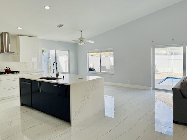 kitchen featuring white gas stovetop, white cabinetry, sink, wall chimney range hood, and a center island with sink