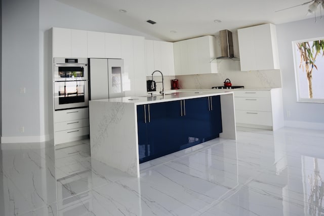 kitchen featuring sink, white cabinetry, a kitchen island with sink, double oven, and wall chimney exhaust hood