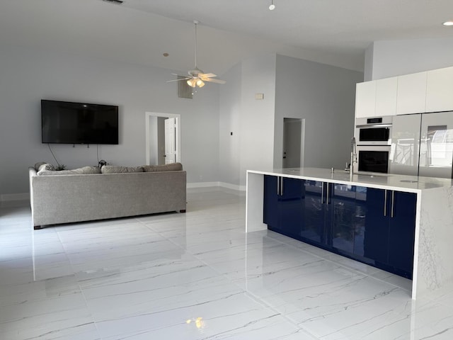 kitchen featuring ceiling fan, white cabinetry, double oven, a towering ceiling, and a center island with sink