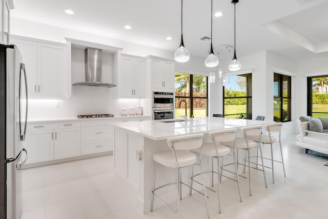 kitchen with white cabinetry, wall chimney range hood, stainless steel appliances, hanging light fixtures, and an island with sink