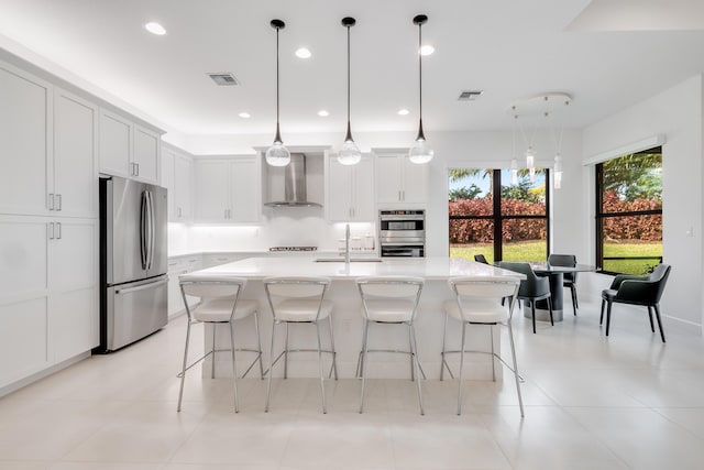 kitchen with a center island with sink, stainless steel appliances, wall chimney range hood, pendant lighting, and white cabinetry