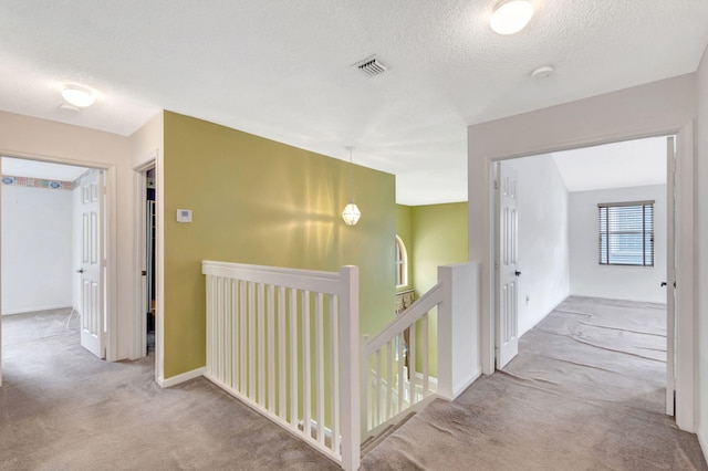 hallway featuring light carpet, visible vents, a textured ceiling, and an upstairs landing