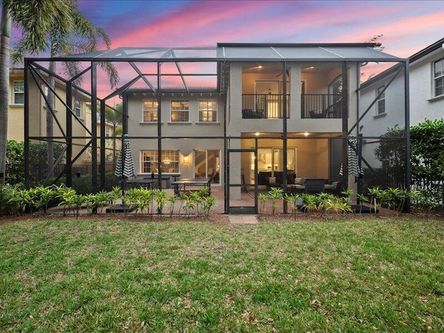 back house at dusk with a balcony, a lanai, a patio, and a lawn