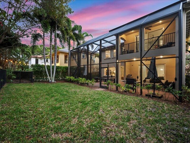 back house at dusk with a lawn, a balcony, and glass enclosure