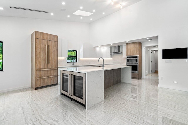 kitchen featuring a skylight, double oven, an island with sink, a towering ceiling, and wall chimney range hood