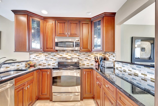 kitchen with appliances with stainless steel finishes, brown cabinetry, glass insert cabinets, a sink, and dark stone countertops