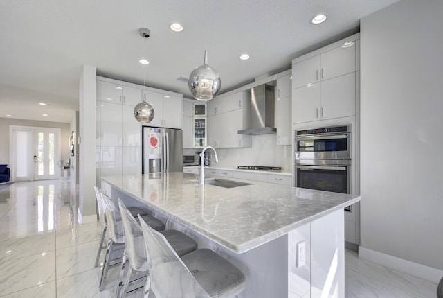 kitchen featuring pendant lighting, sink, wall chimney range hood, appliances with stainless steel finishes, and a center island with sink