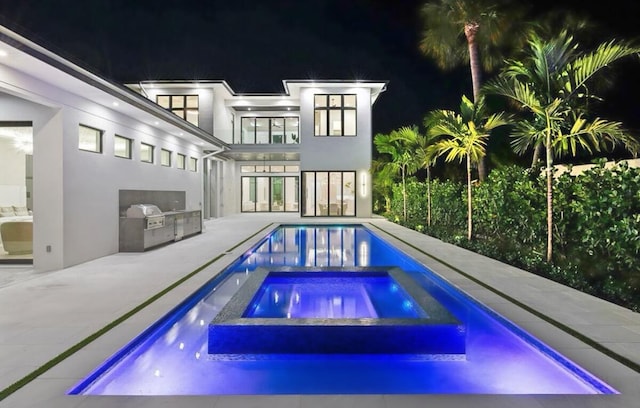 pool at night with an in ground hot tub, an outdoor kitchen, and a patio area