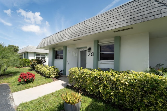 view of exterior entry featuring a yard, a shingled roof, mansard roof, and stucco siding